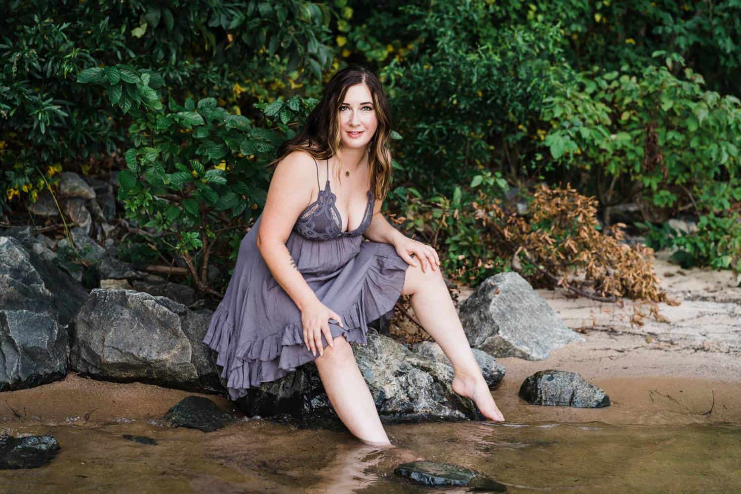 A woman in a short purple nightie sits at the edge of a creek with her feet in the water