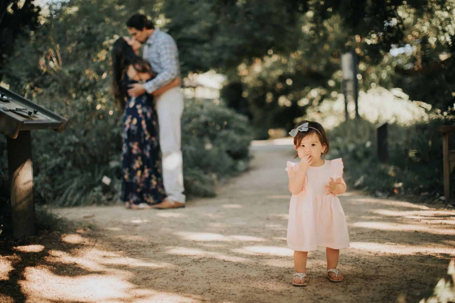 Two parents kiss in the background. In the foreground, their toddler daughter stands in a pink dress looking solemnly at the camera.