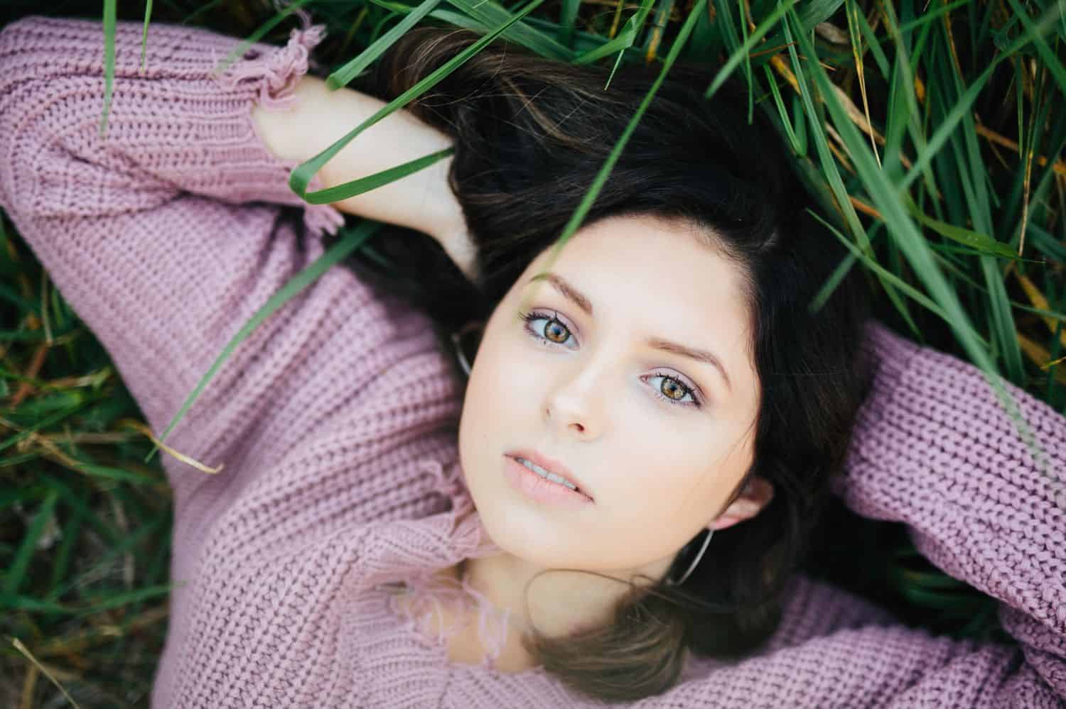A high school senior in a lavender sweater lies in the green grass with a serious expression on her face