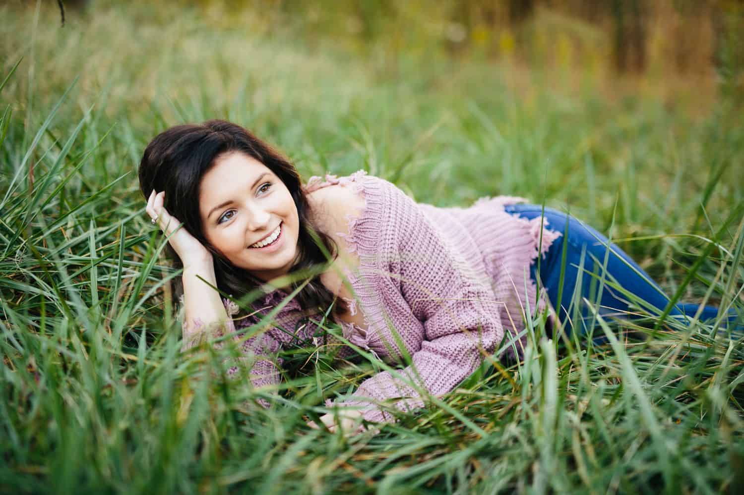 A high school senior wearing a lavender sweater lies on her side in long, green grass