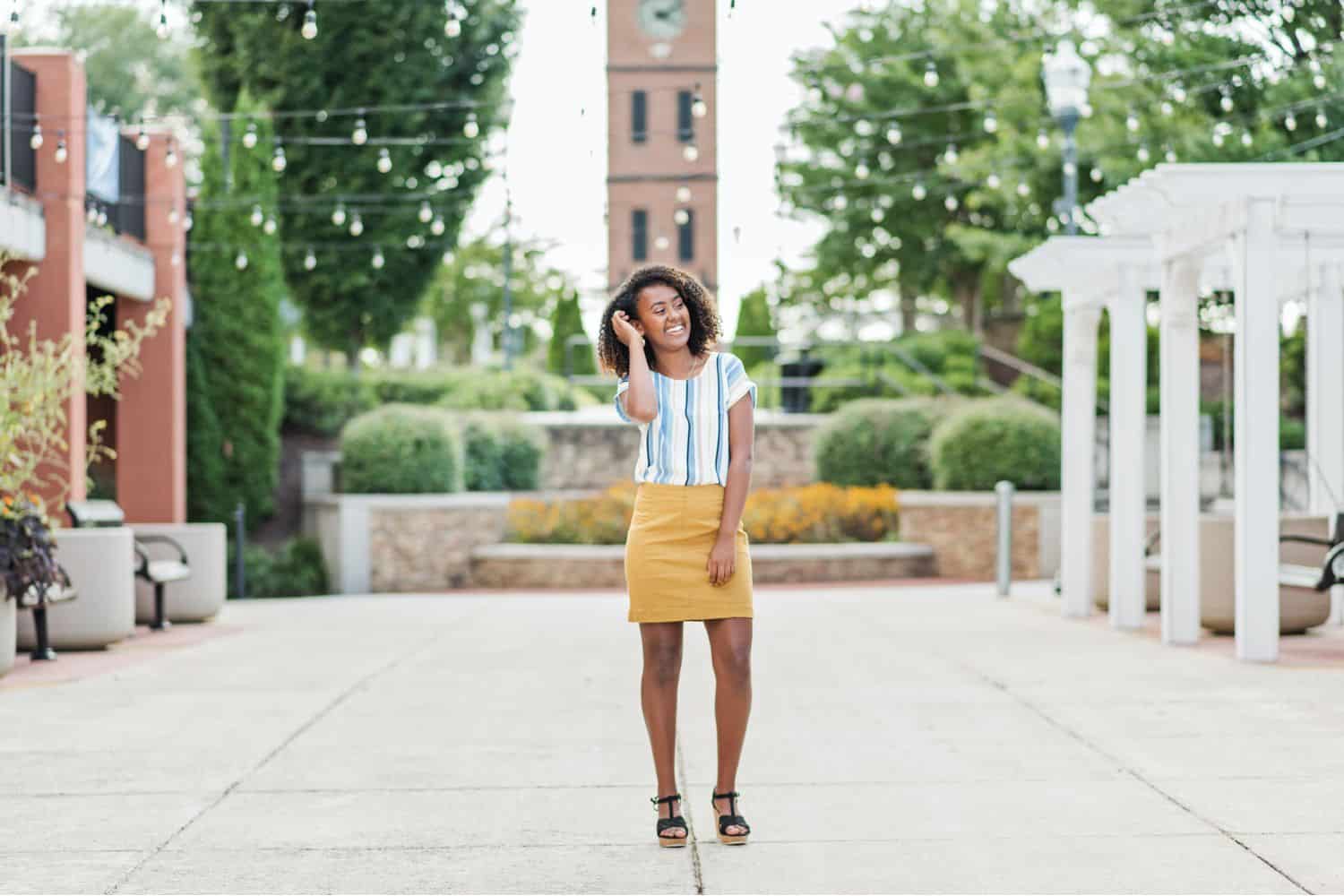A high school senior girl poses in the town square wearing a mustard yellow mini skirt and a blue-striped white blouse.