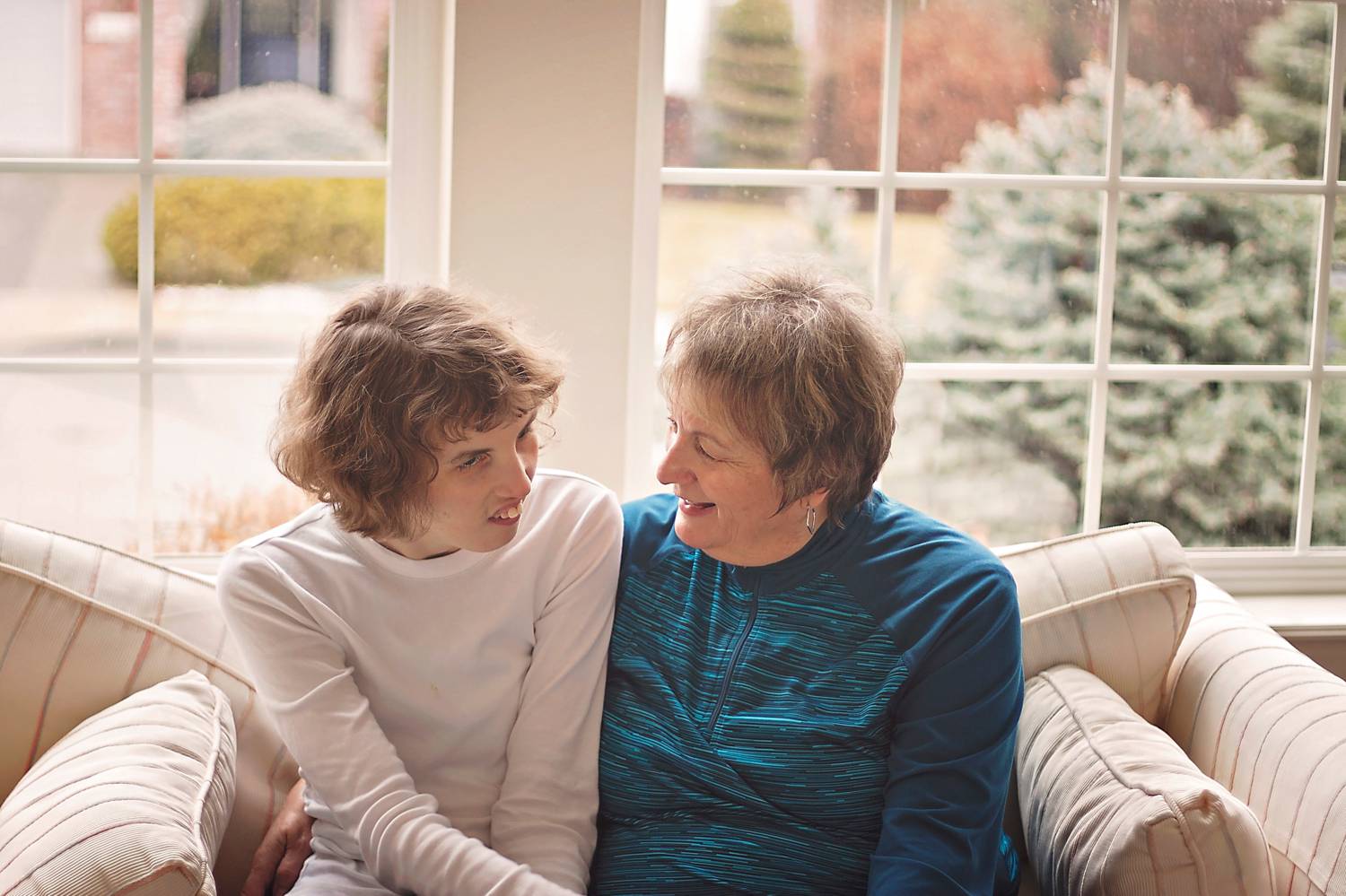 A mother and daughter sit closely on a sofa at home, sharing an intimate moment.