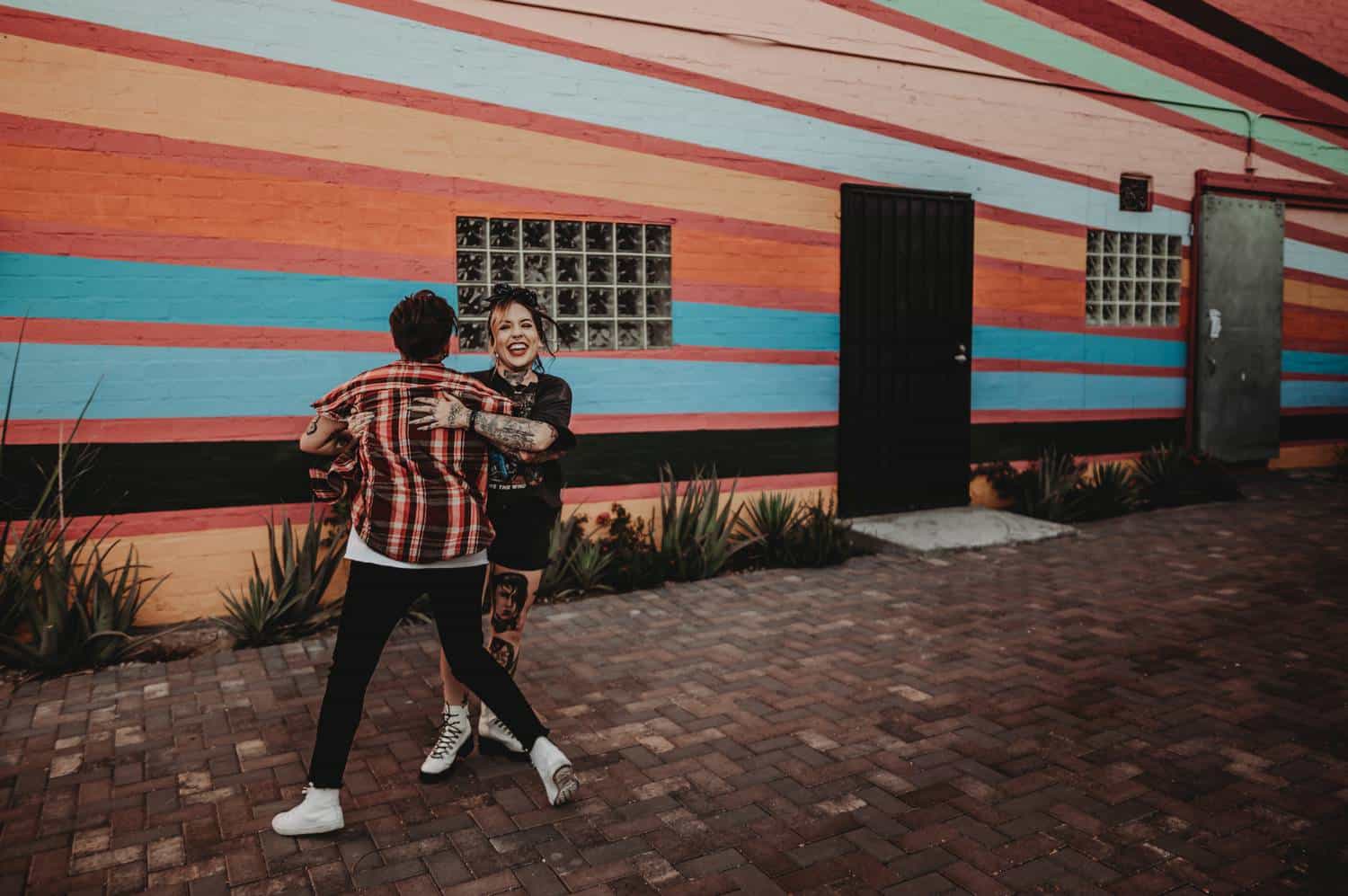 A couple dances joyfully on a stone-paved pathway into a rainbow-striped building.