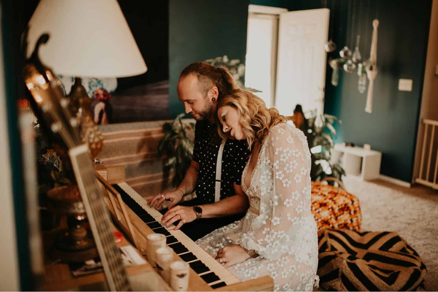 A couple sits at a piano in their eclectically decorated home. The woman's head rests on the man's shoulder as he plays. Learn how to take low light photos like this one by Shelby Laine Photography.