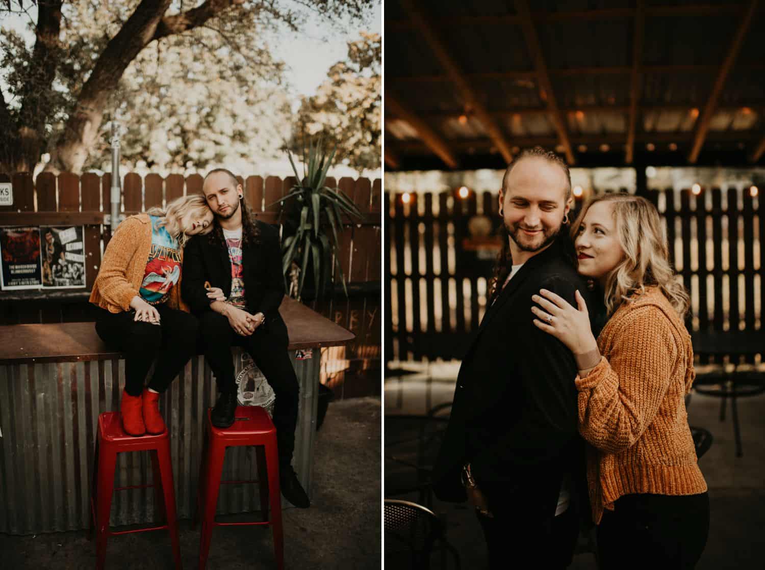 A hipster man and woman sit on red barstools at an outdoor bar surrounded by a picket fence.