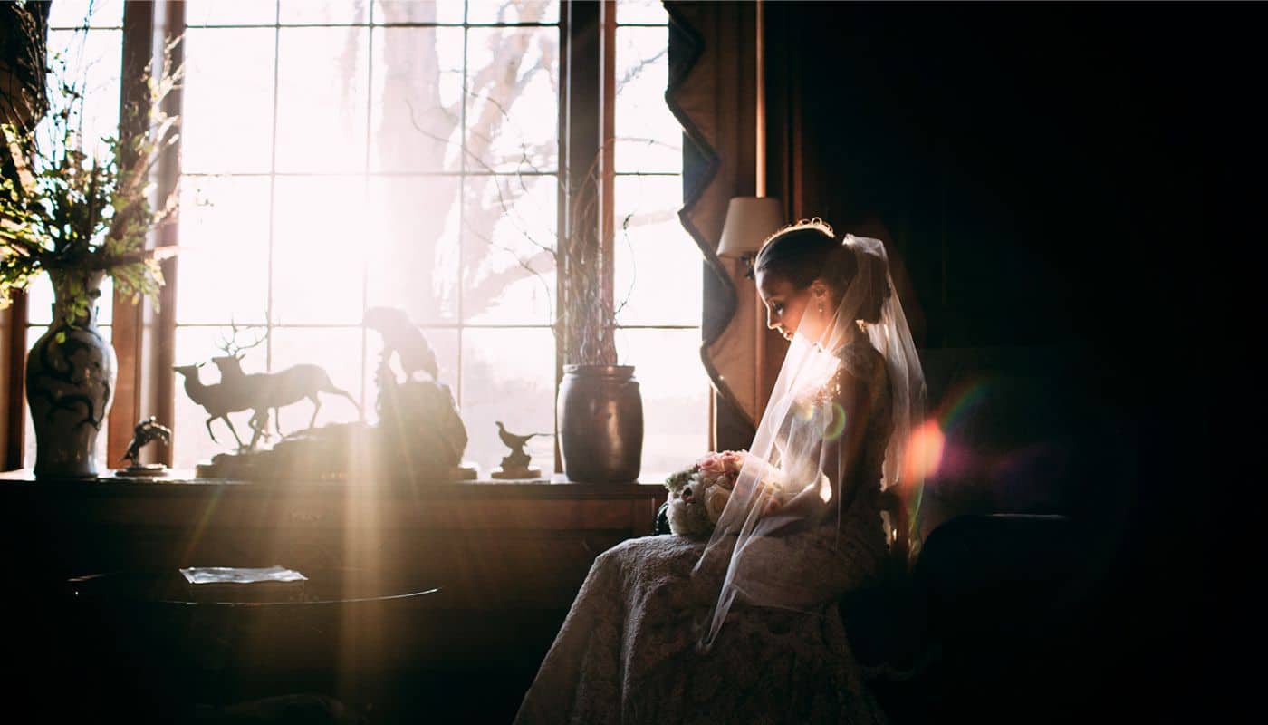 A bride in a long veil is backlit by the sun streaming through a large window in this wedding photo shot by photographer Ginny Corbett