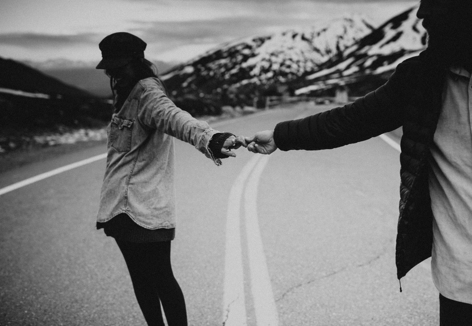 A woman leads a man across an empty mountain road with snow in the background.