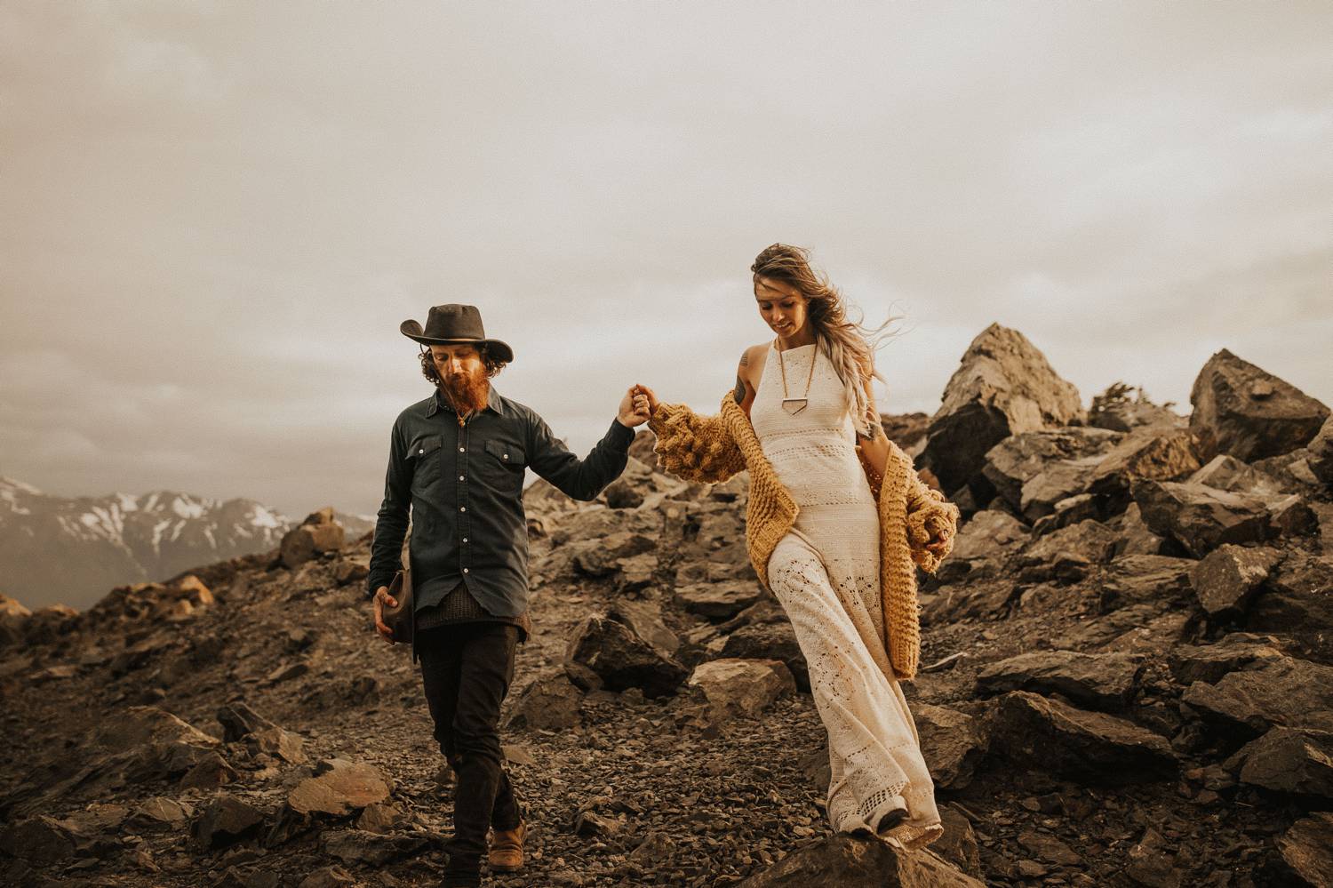 A man in a black hat holds the hand of a woman in white as they walk across a rocky beach