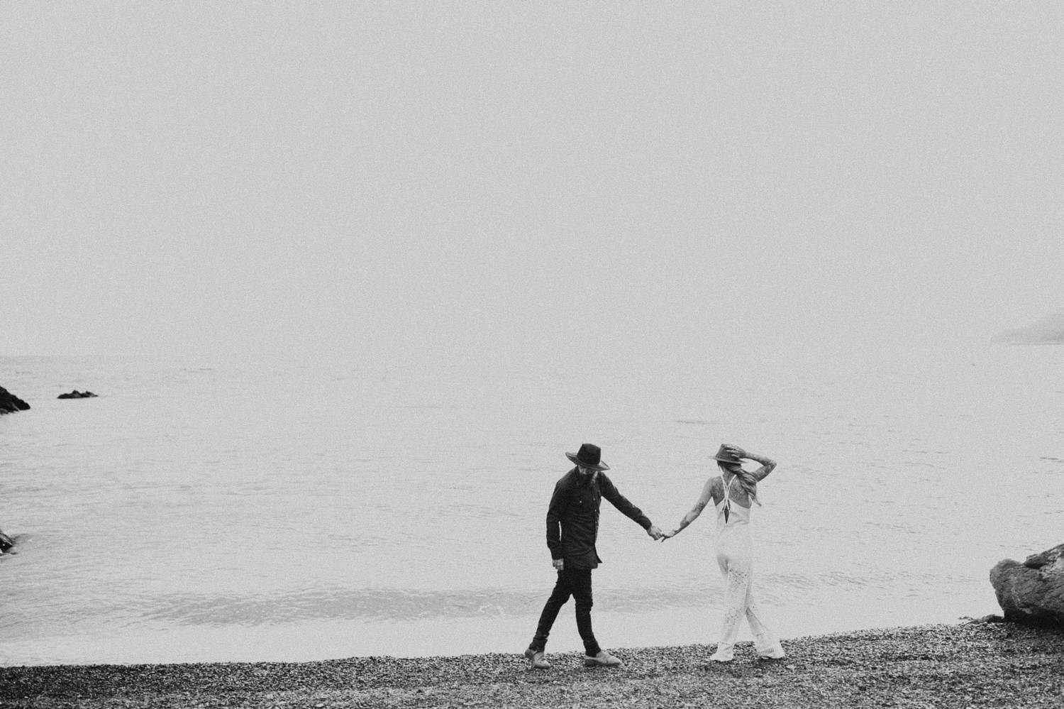 A man dressed in black holds hands with a woman dressed in white as they stroll along the beach in this moody black and white photo by Brittany Boote