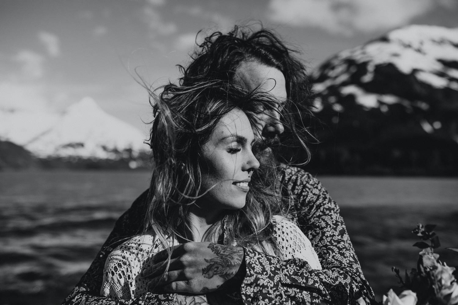 A man stands behind a woman and wraps his arms lovingly around her. Their faces are both angled toward the sun