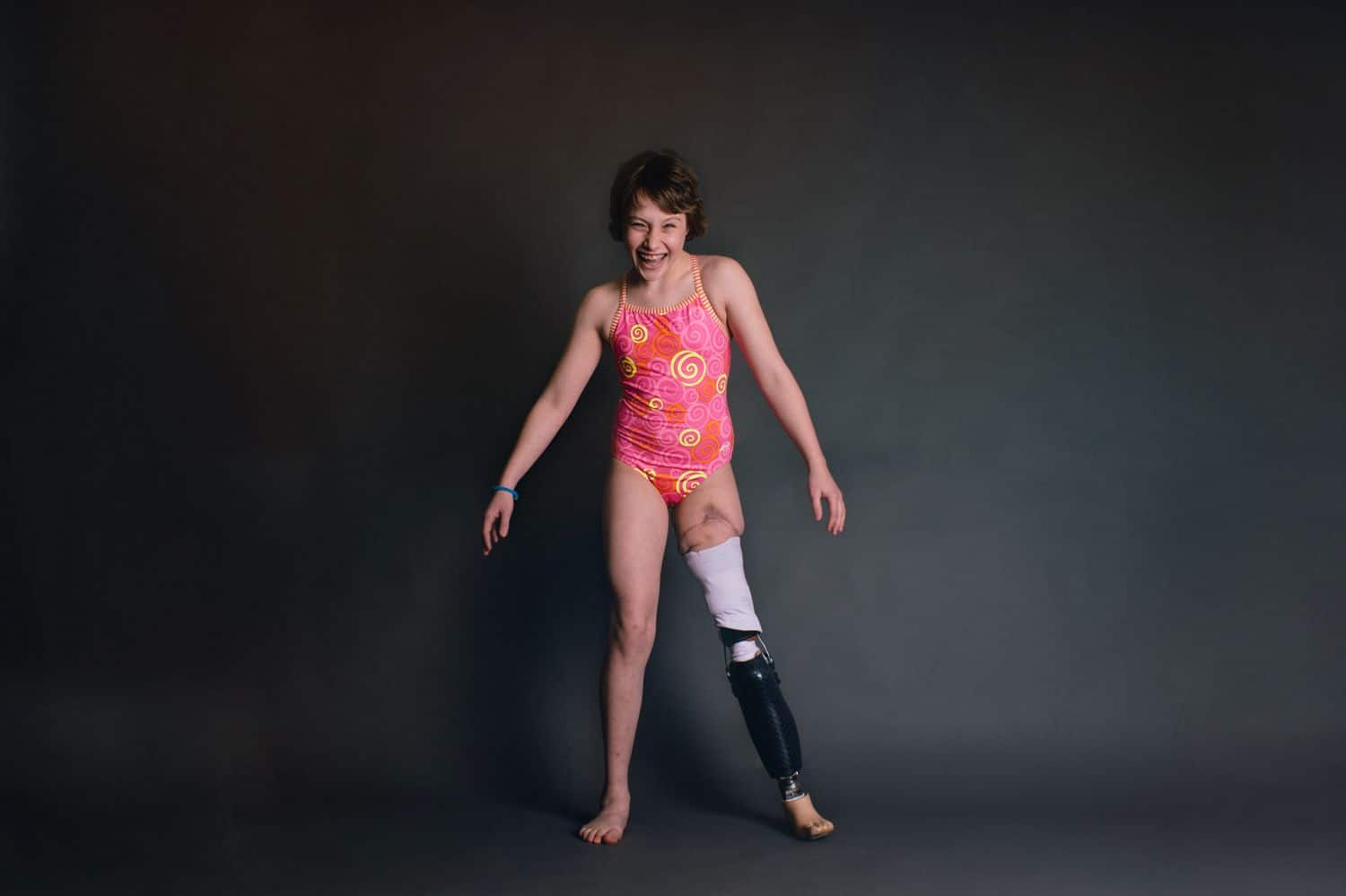A young woman with a rotationplasty poses on a black background while smiling and wearing a pink leotard. By Kate T. Parker for Strong is the New Pretty