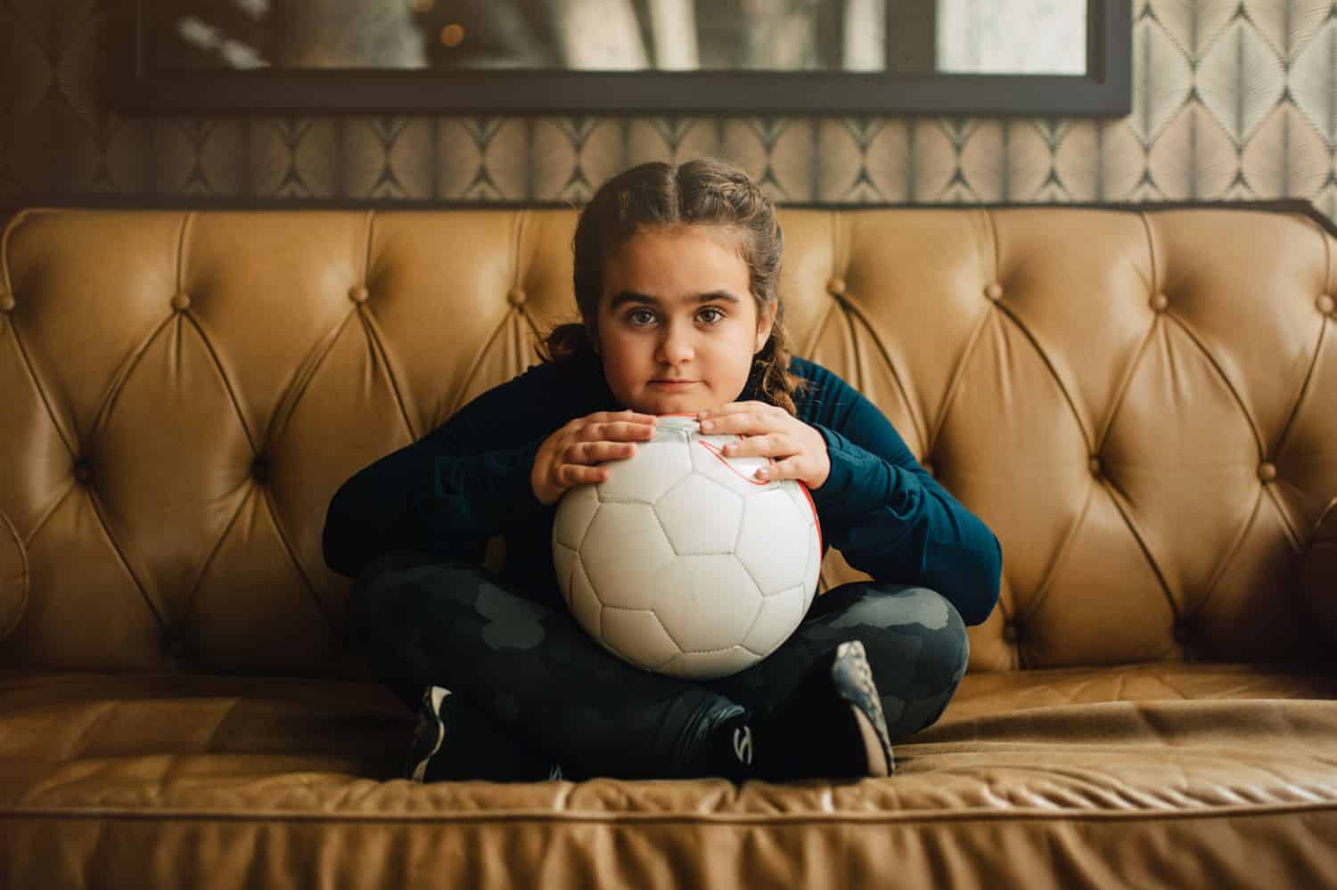 A little girl with French braids sits cross-legged on a leather sofa holding a soccer ball in her lap. By Kate T. Parker for Strong is the New Pretty