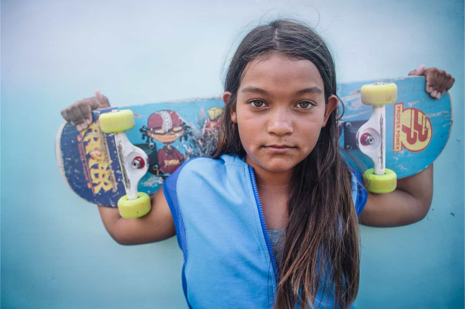 A girl with long messy hair holds a skateboard across her shoulders and gazes directly into the camera. By Kate T. Parker for Strong is the New Pretty
