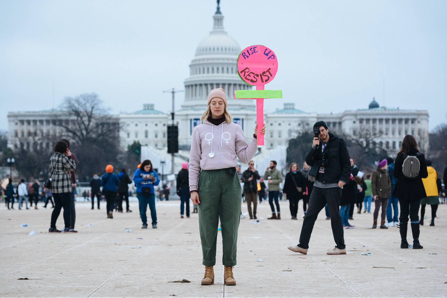 Photo: Kate Warren's documentary image of a young, white, blonde woman in a pink hat and jacket. She stands in front of the U.S. Capital holding a sign that says, "Rise up. Resist."