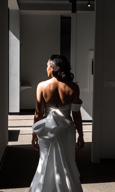 A woman in a white strapless gown with a large bow in the back walks down a hallway with light streaming in. She is wearing a jeweled headpiece and earrings.
