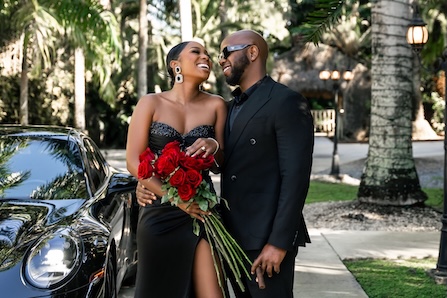 A stylish couple, dressed in all black, stands affectionately next to a sleek black car, with the woman holding a bouquet of red roses. They are both smiling and looking at each other, set against a backdrop of lush greenery and palm trees.