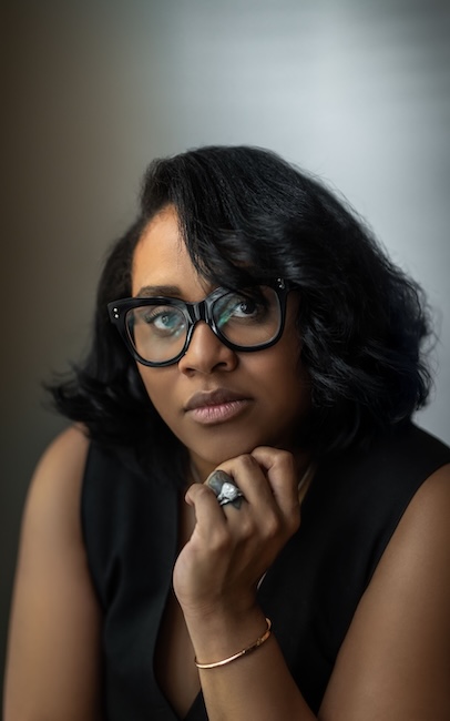A portrait of a woman with dark hair and glasses, wearing a black sleeveless top, with her hand resting on her chin. She is wearing a large ring and a bracelet.