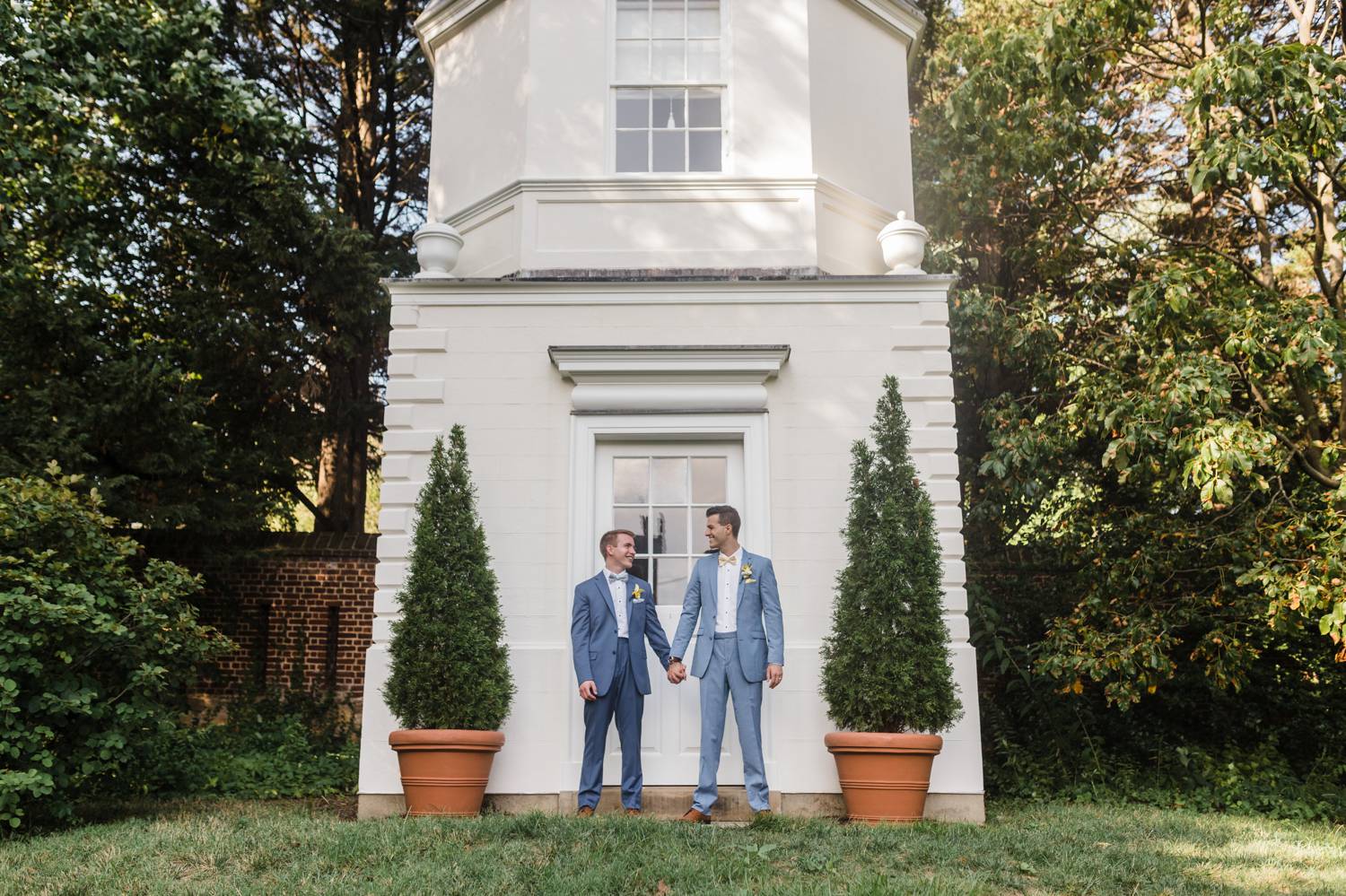 Two grooms dressed in gray suits hold hands in front of a tall white chapel