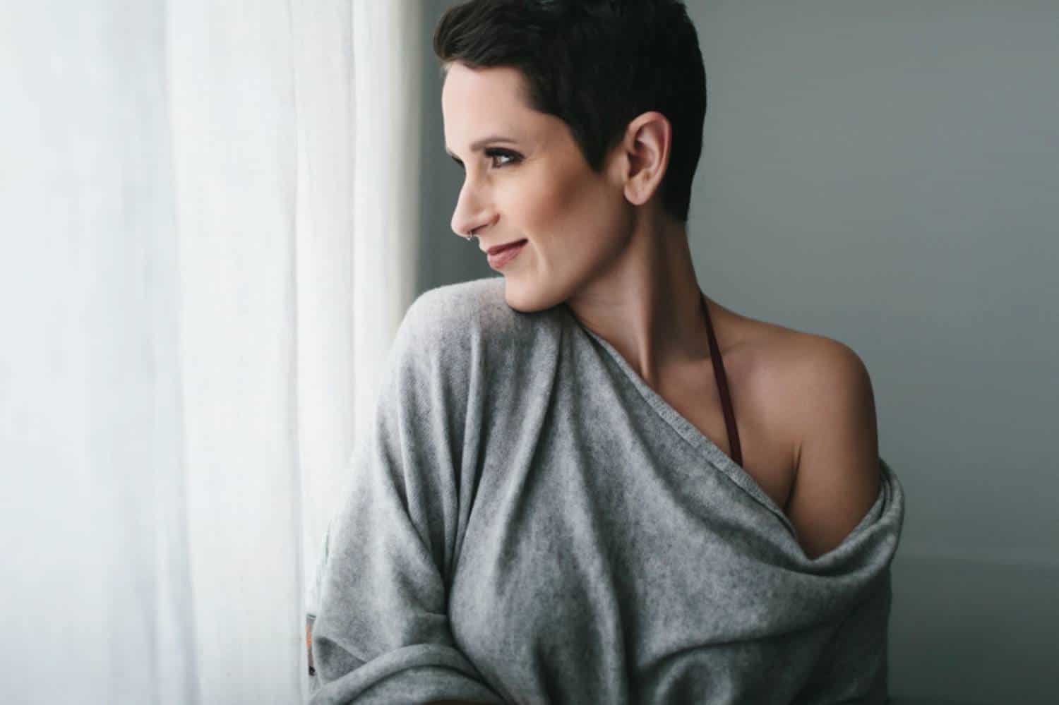 A woman with a brunette pixie cut, wearing a gray cashmere sweater that has slipped off one shoulder, looks over her shoulder out the window with a slight smile on her face. Learn how to take boudoir photos for clothed clients by reading the ShootProof blog!