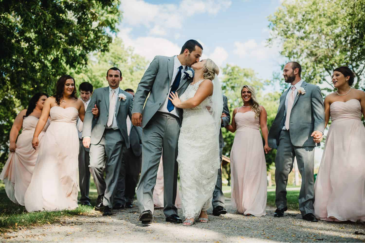 A bride and groom kiss in the middle of the street as their wedding party walks behind them.