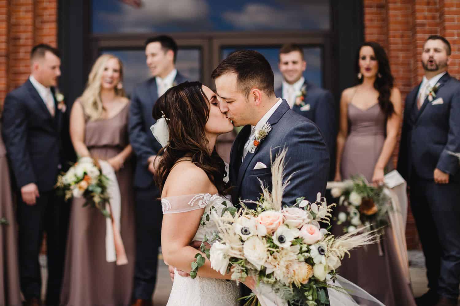 A bride and groom kiss as their wedding party stands in the background.
