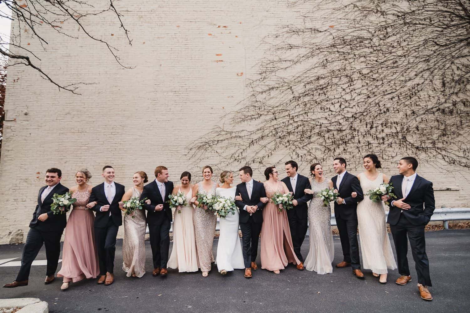 A wedding party in colorful dresses and black tuxes poses in front of a vine-covered wall.