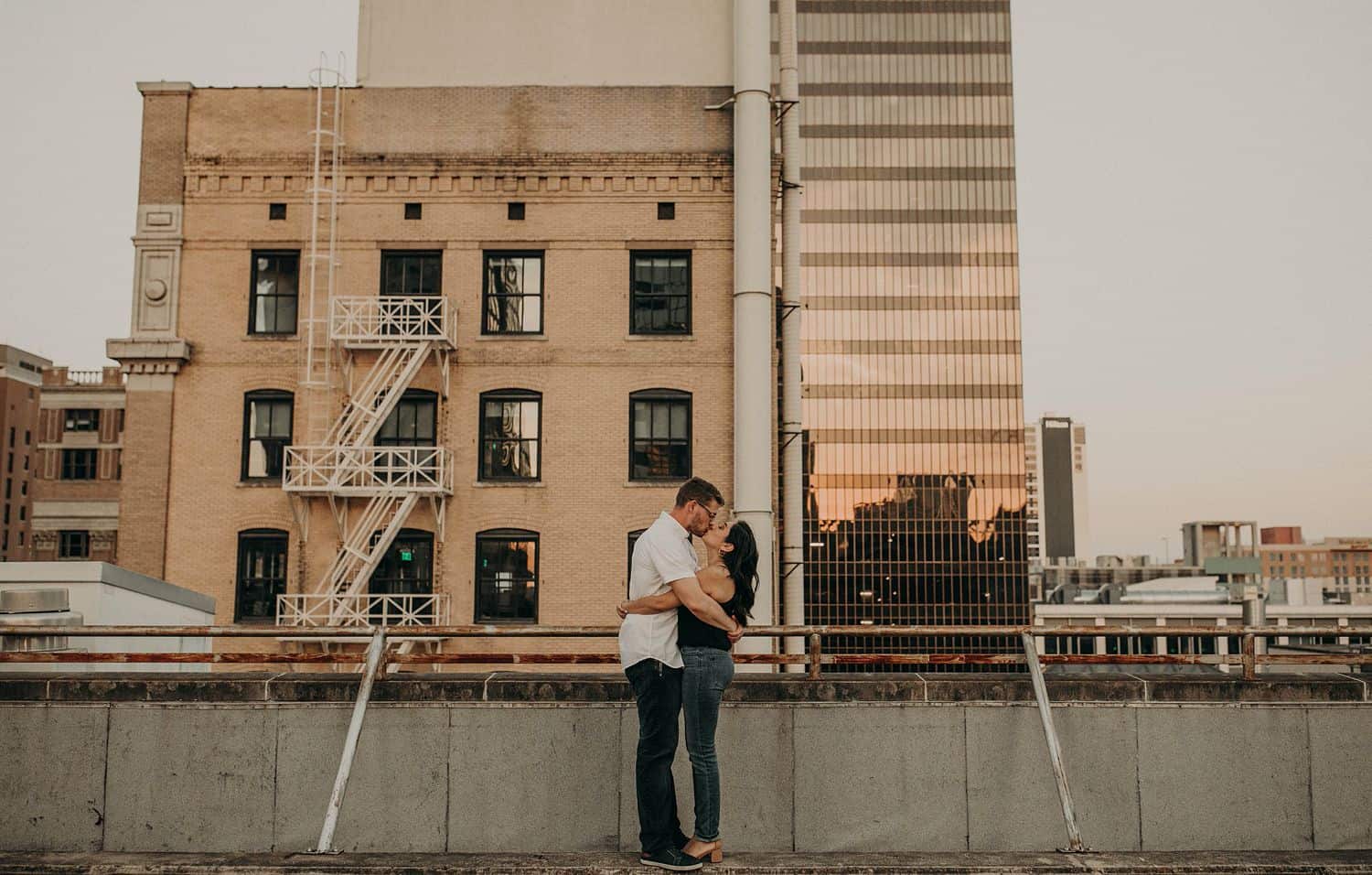 A man and woman hug and kiss on a rooftop overlooking a brick building with a fire escape