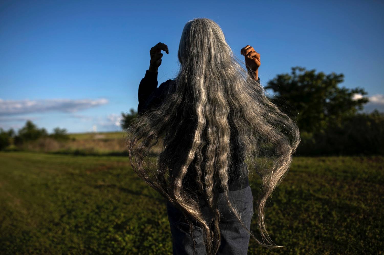 Photo: Maddie McGarvey's portrait of a woman with flowing grey hair shows her brushing her hair back so it flows below her waist as she looks out at a bright blue sky.