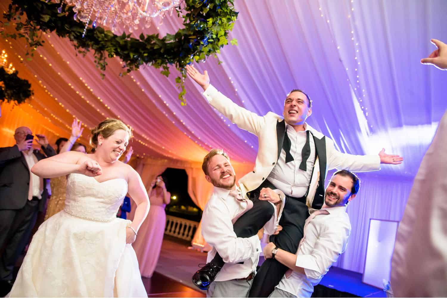 Make Money with Photography: Bride dances while two wedding guests lift Groom into the air.
