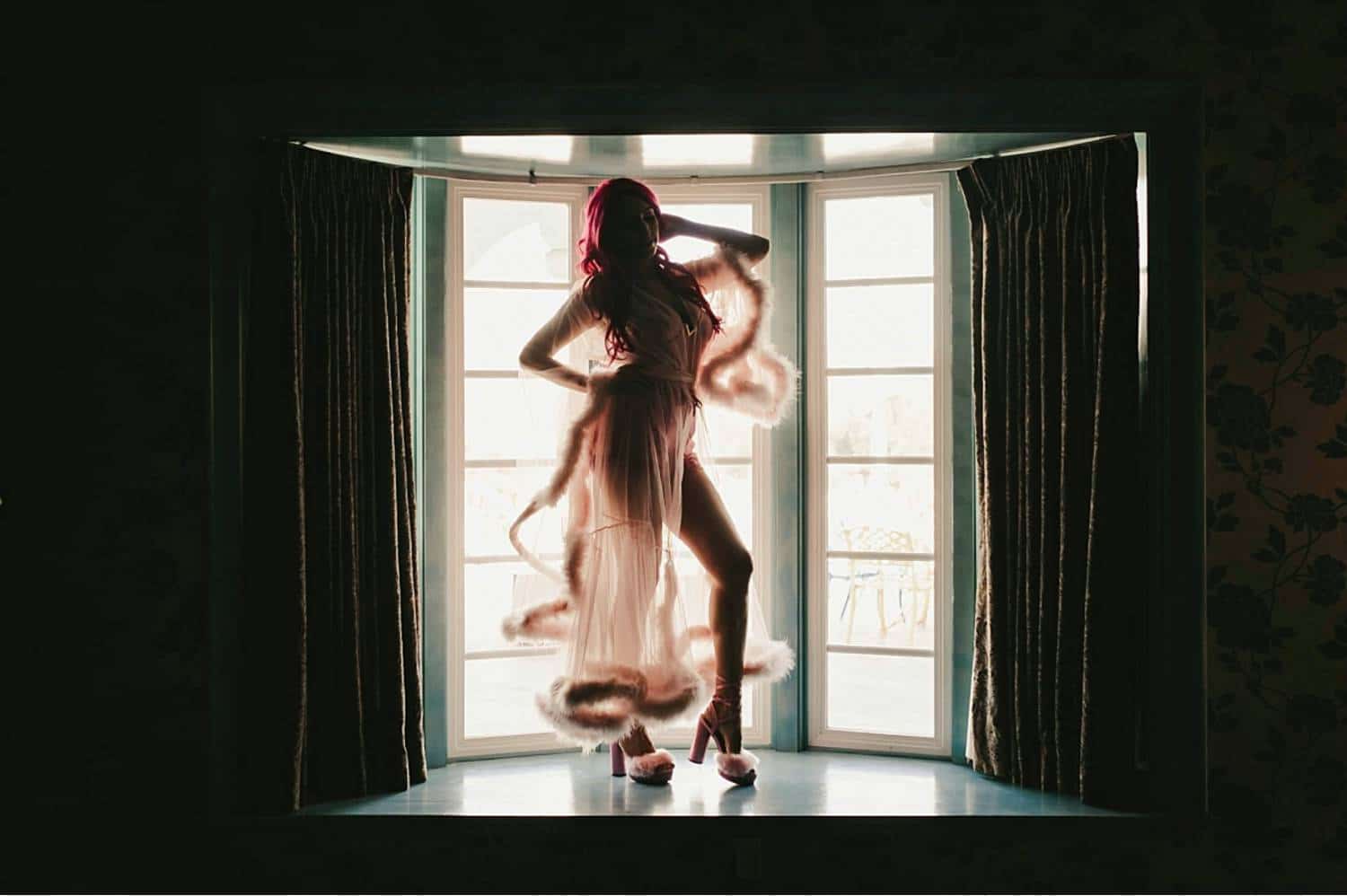 Photo: Michelle Roller's boudoir portrait of a pink-haired woman wearing a silk and fur robe as she stands on a platform before a large bay window.