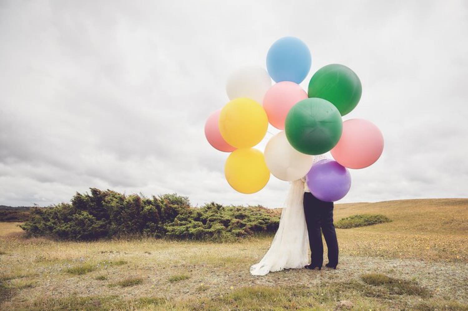 Photo: Mille Stengaard's portrait of a bride and groom is shot against a plane, grey sky. The couple holds a massive bunch of colorful balloons that hides their faces.