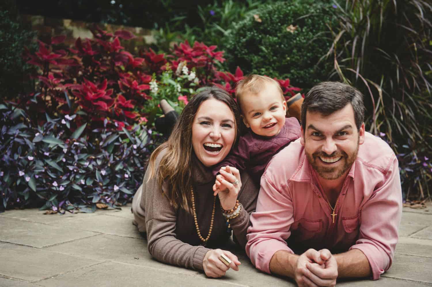 Mom, dad, and baby pose for a photograph while lying on their stomachs in front of a garden of flowers.