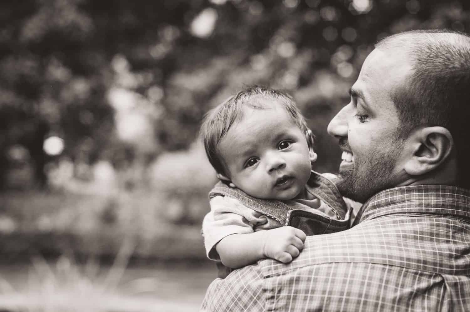 A dad snuggles his toddler son close to his face in black and white.