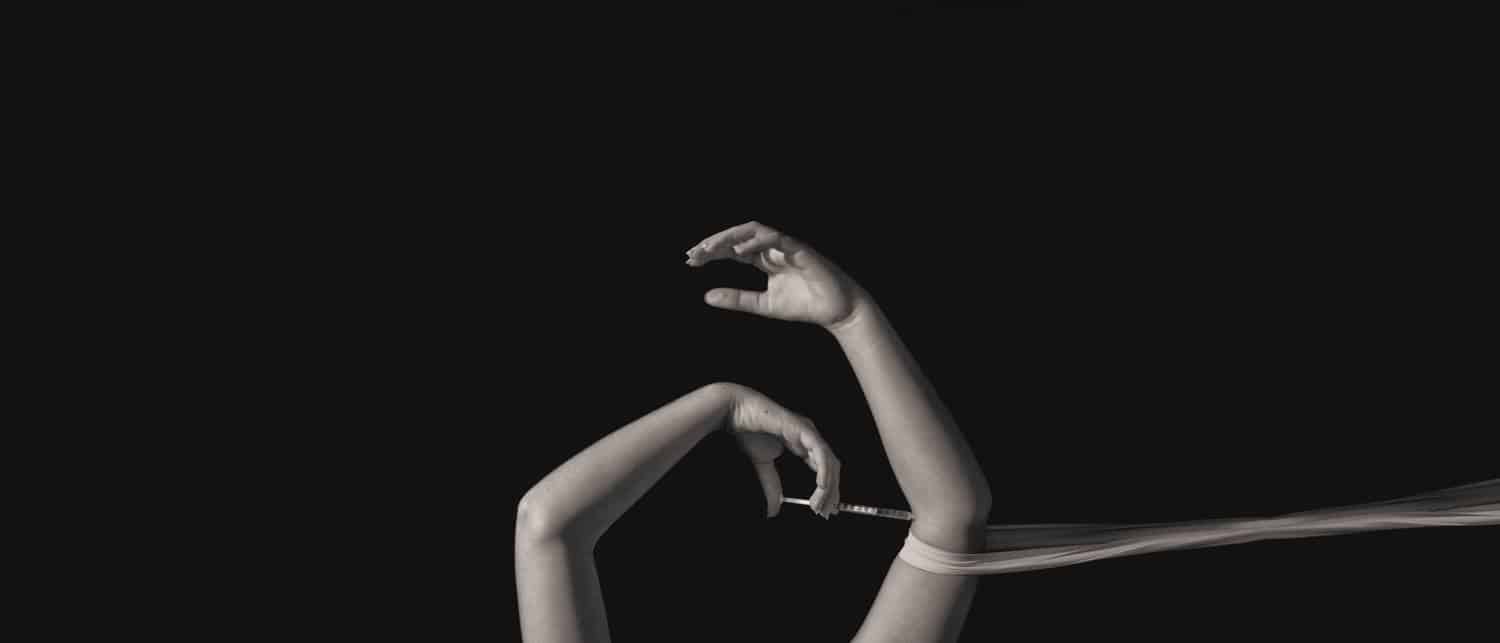 A girl's arms seem to float against a black background. One hand relaxes open as a scarf tightens around the elbow. The other hand holds a syringe and appears to be injecting a substance into the other arm.