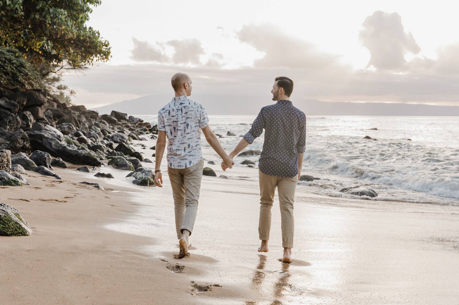 Two men stroll hand-in-hand on the beach at sunset.