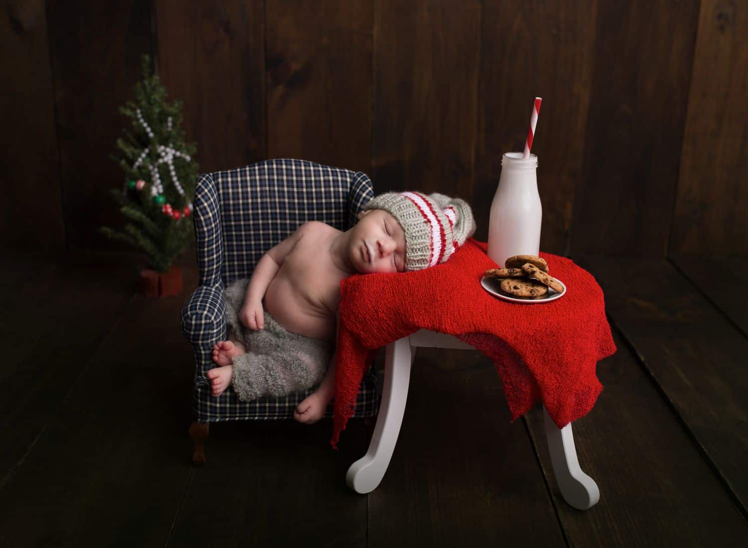 A tiny baby is posed with newborn photography props: a tiny chair and table topped with a red cloth, a bottle of milk, and a plate of miniature cookies.