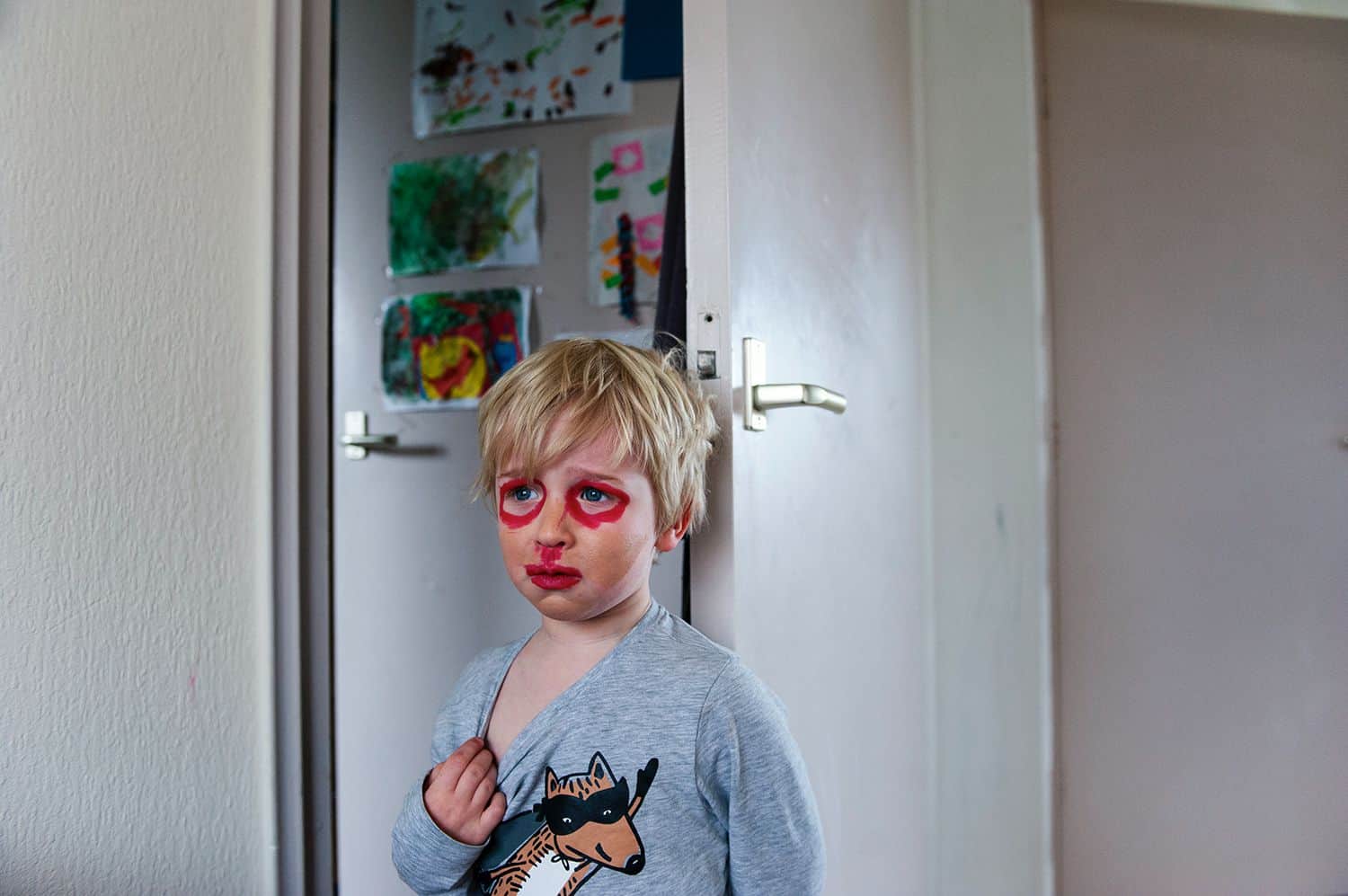 A little boy tugs at the neck of his sweatshirt looking distressed as he stands in a kitchen wearing self-applied makeup