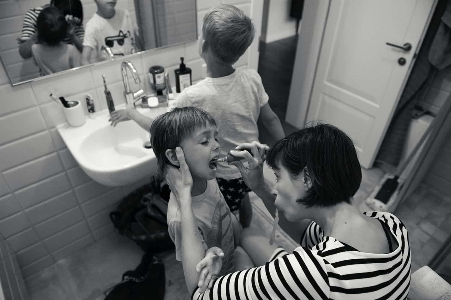 A mom brushes her younger child's teeth while her older child rinses their mouth at the sink