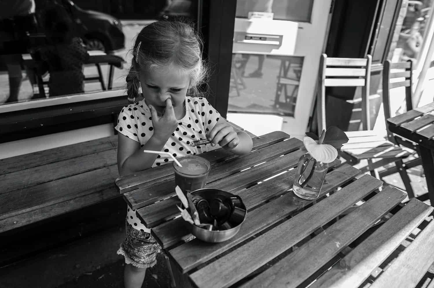 A little girl with a braid sits at an outdoor dining table picking her nose