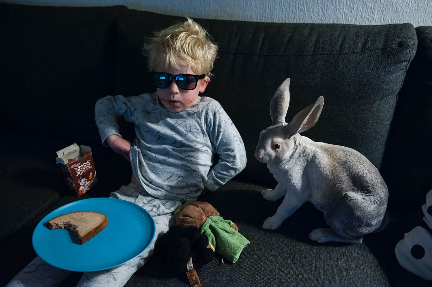 A little boy in sunglasses sits on a sofa eating a piece of bread with a rabbit statue sitting beside him
