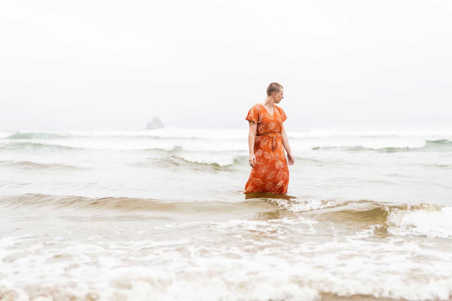 A person with a shaved head wearing a rust-colored dress stands in the ocean waves while photographer Sofia Angelina conducts a healing session that addresses chronic pain.