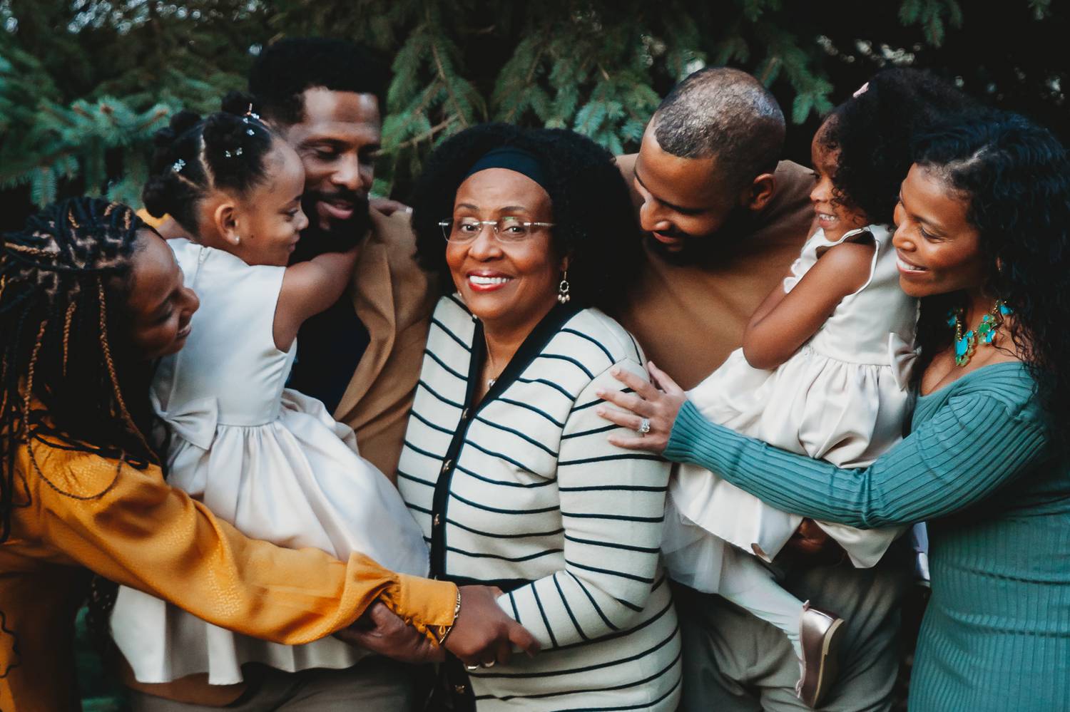 A smiling Black family gathers around the matriarch and hugs one another close