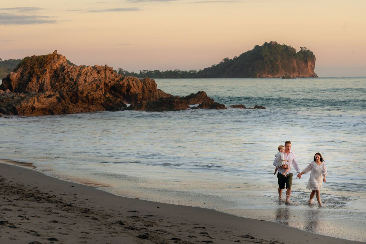 Client Experience: A photo by Kevin Heslin depicts a couple standing in the surf of a beach at sunset while holding their young child.