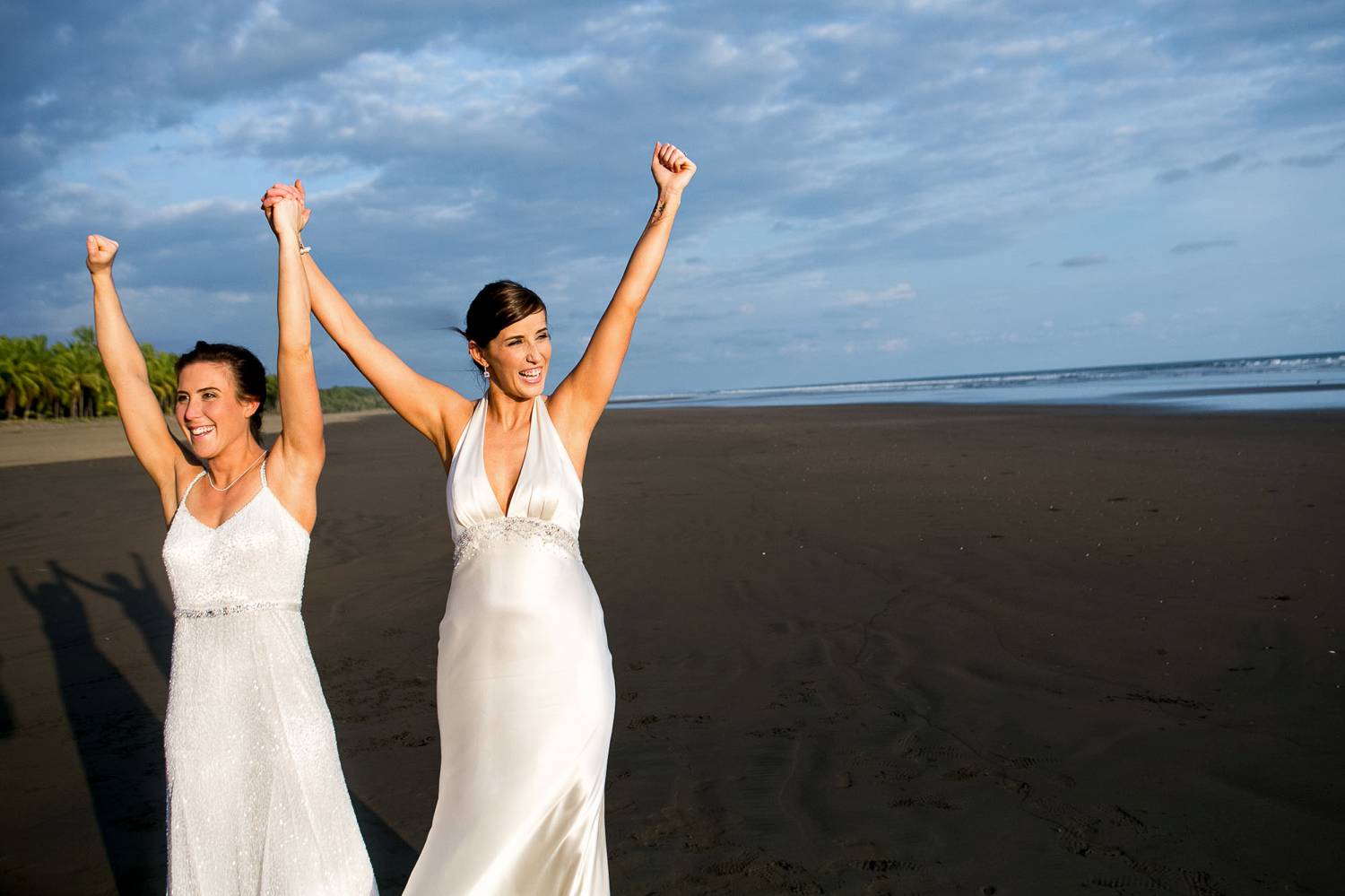 Client Experience: A wide-angle photo by Kevin Heslin depicts two brides in white silk dresses with their arms raised to the sky on the beach.