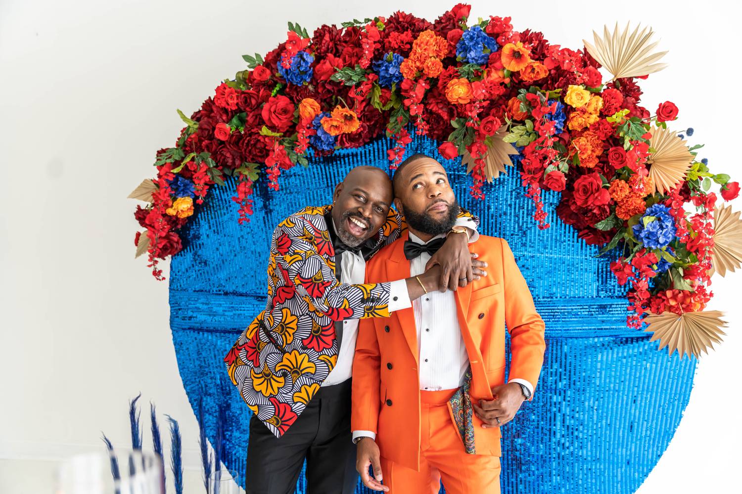A groom in a colorful jacket wraps his arms around the shoulders of his husband who wears a bold orange suit