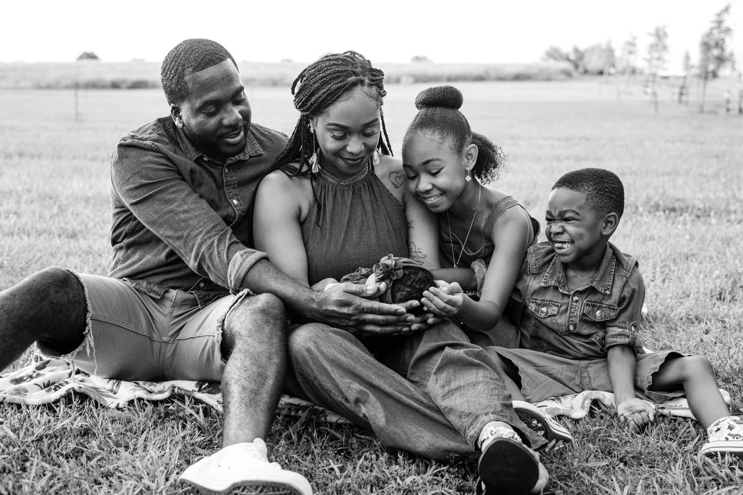 A smiling Black family sits on a quilt in a field with their hands collectively cradling the family's new baby.