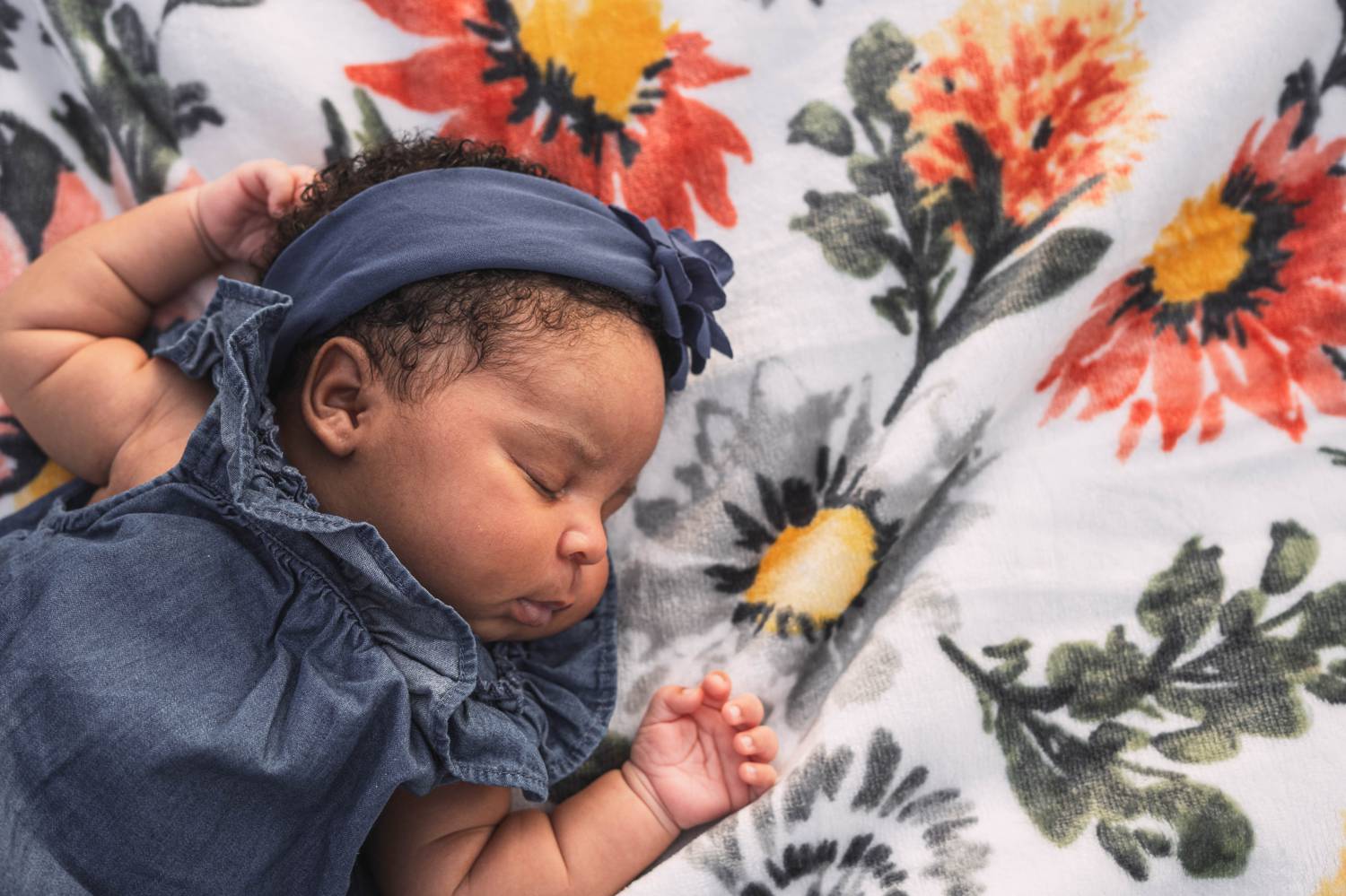 A newborn baby in a ruffled denim dress sleeps soundly on her back atop a colorful quilt.