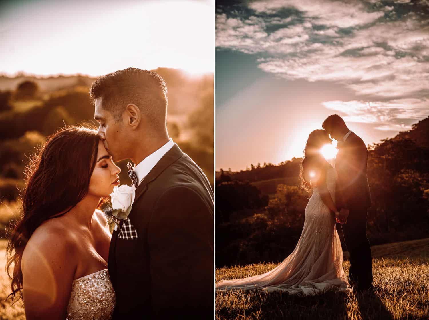 Groom kisses bride’s forehead with sun setting behind them. Silhouette of bride and groom standing face to face with sun shining between their bowed heads.