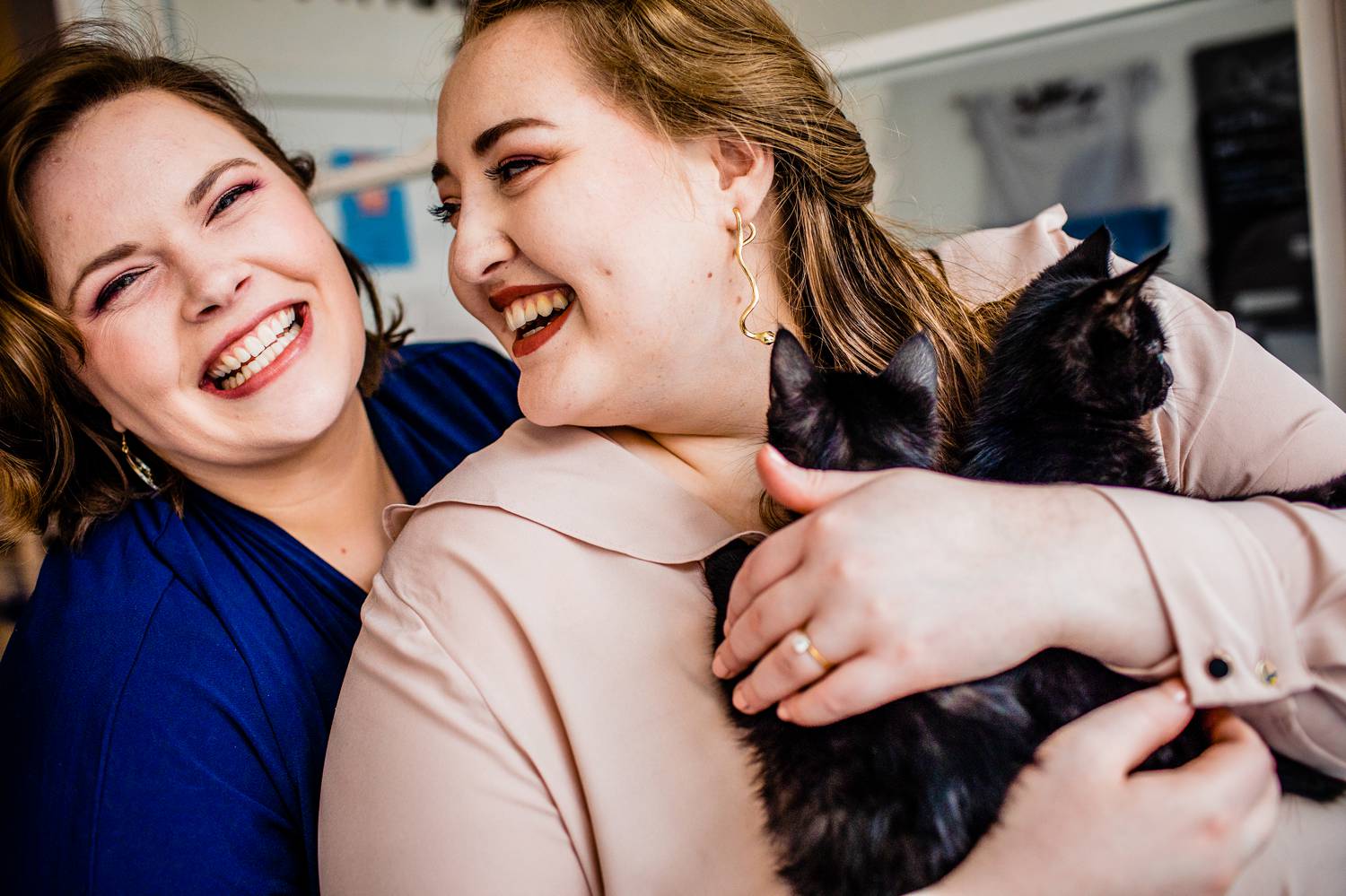 A woman in a blue shirt smiles at the camera over the shoulder of her wife, who sits in front wearing a peach shirt and holding two kittens