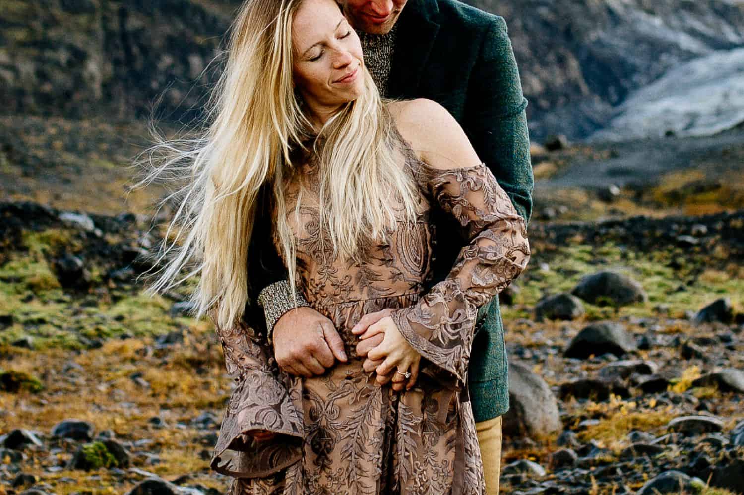 Woman in a brown lace dress is embraced from behind by a man in a green velvet jacket. Only the woman's face is shown.